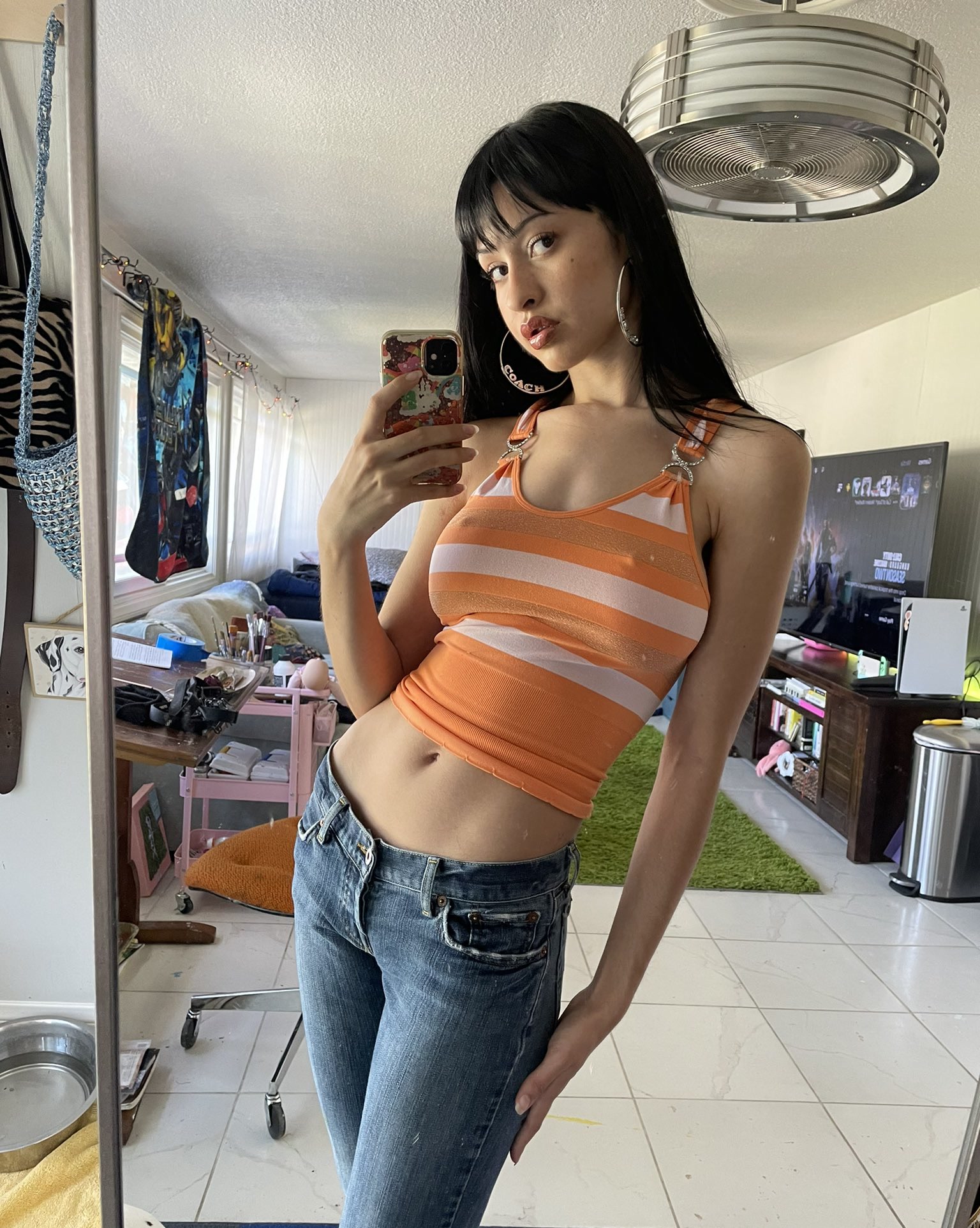 Mirror selfie with long dark hair, an orange striped halter top and jeans in a sunlit bedroom.